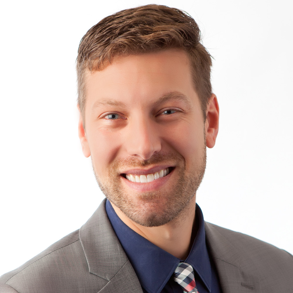 Headshot of a brown haired male who is smiling and has a light beard. His suit coat is gray and he is wearing a blue shirt with a blue, white and red plaid tie.