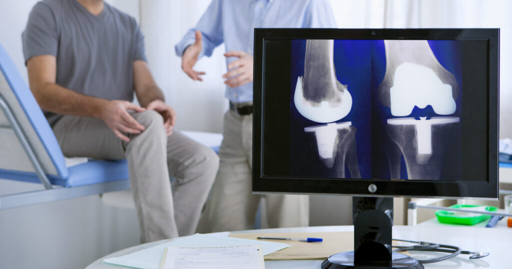 Computer monitor displaying a knee replacement xray in the foreground. Patient and doctor in the background.