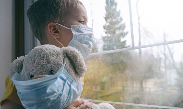 Young boy wearing mask, holding teddy bear, looking out the window