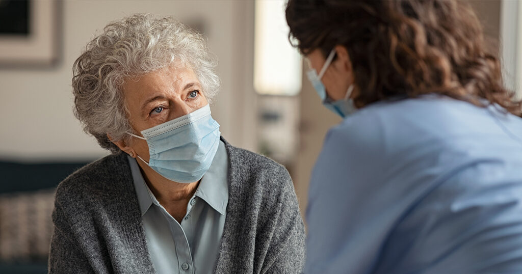 Older female patient talking to her healthcare provider, both wearing masks