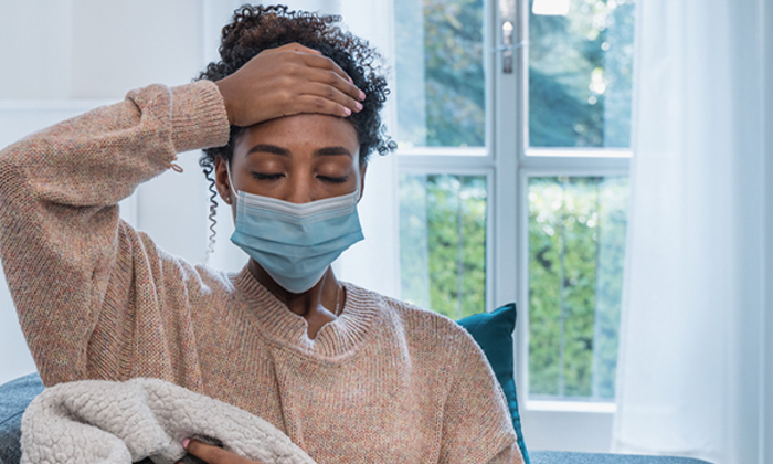 Woman wearing face mask sitting under a blanket and holding forehead,