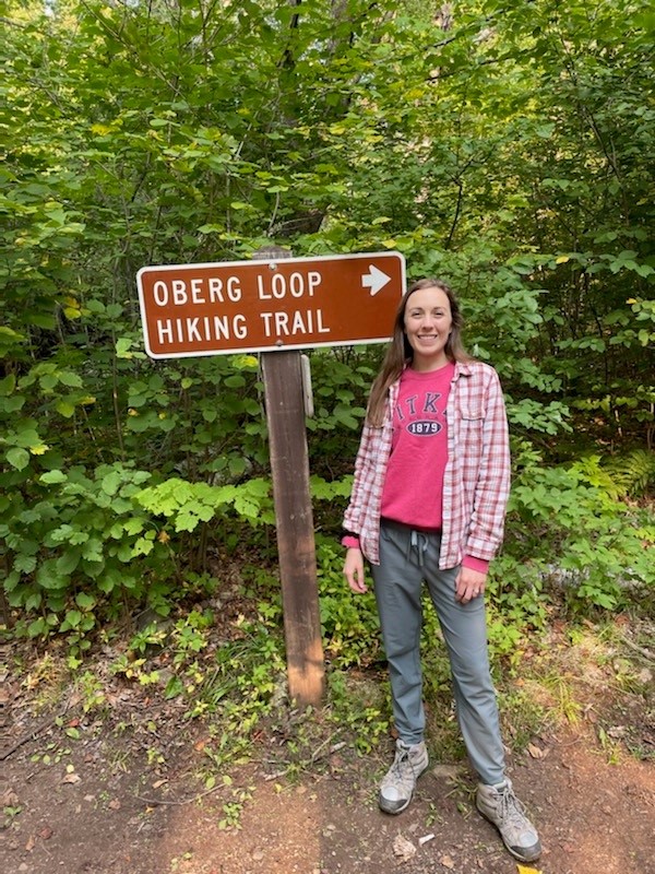 Young lady with long brown hair in red flannel shirt standing next to a sign. 