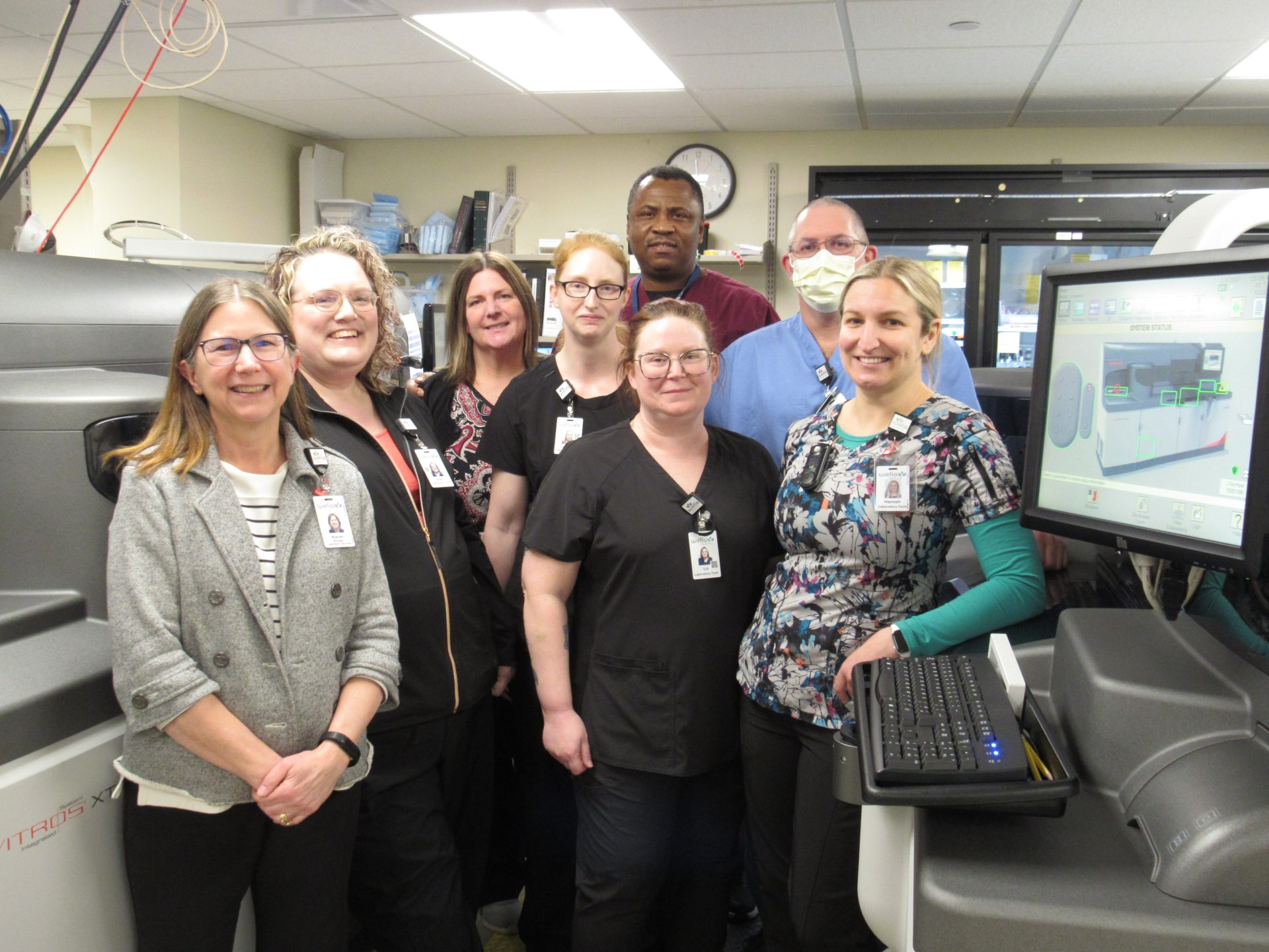 Welia Health laboratory employees standing between the 2 new lab analyzers at the mora campus, smiling.