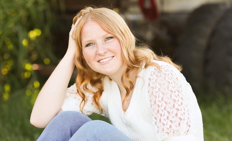 Smiling blondish-red colored young lady posed sitting with elbow on knee and hand on the side of her head.