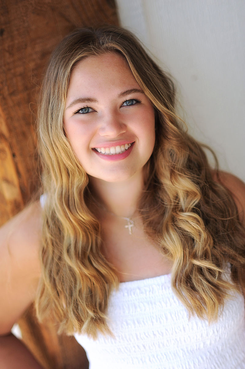 Young lady smiling with long blondish-brown hair wearing a Christian cross necklace and smiling with her teeth showing.