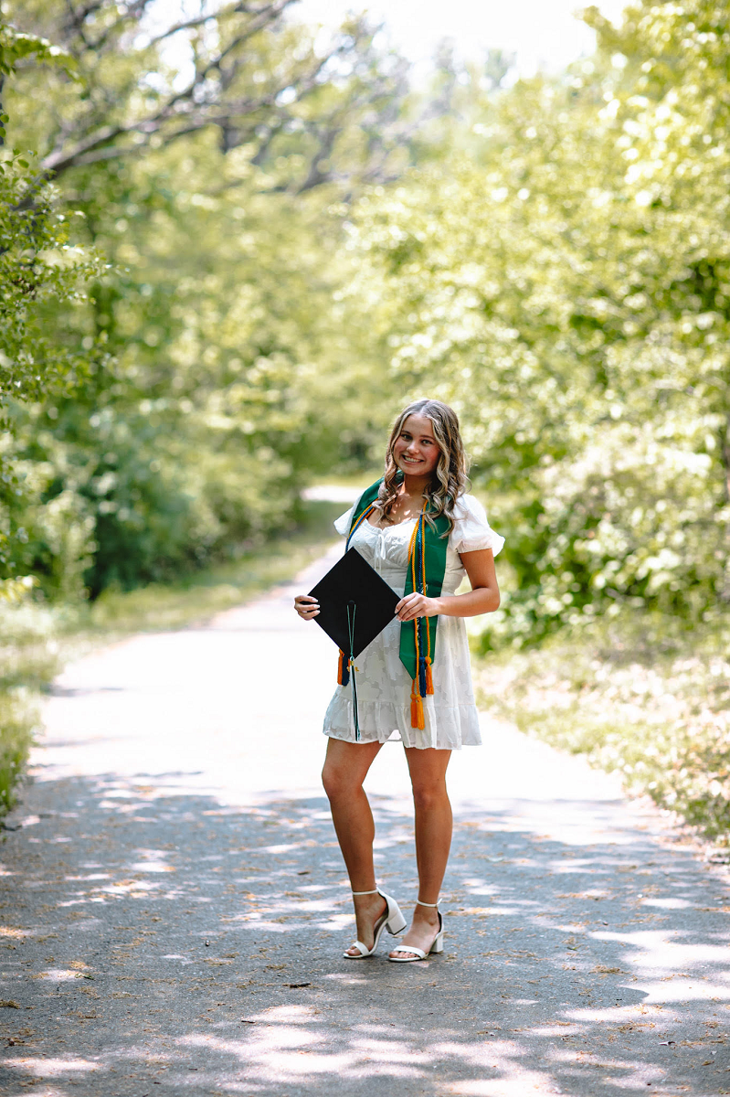 Young lady smiling with blondish-brown hair wearing a white dress and graduation cords, holding her graduation cap while standing on a tarred bike path surrounded by trees. 