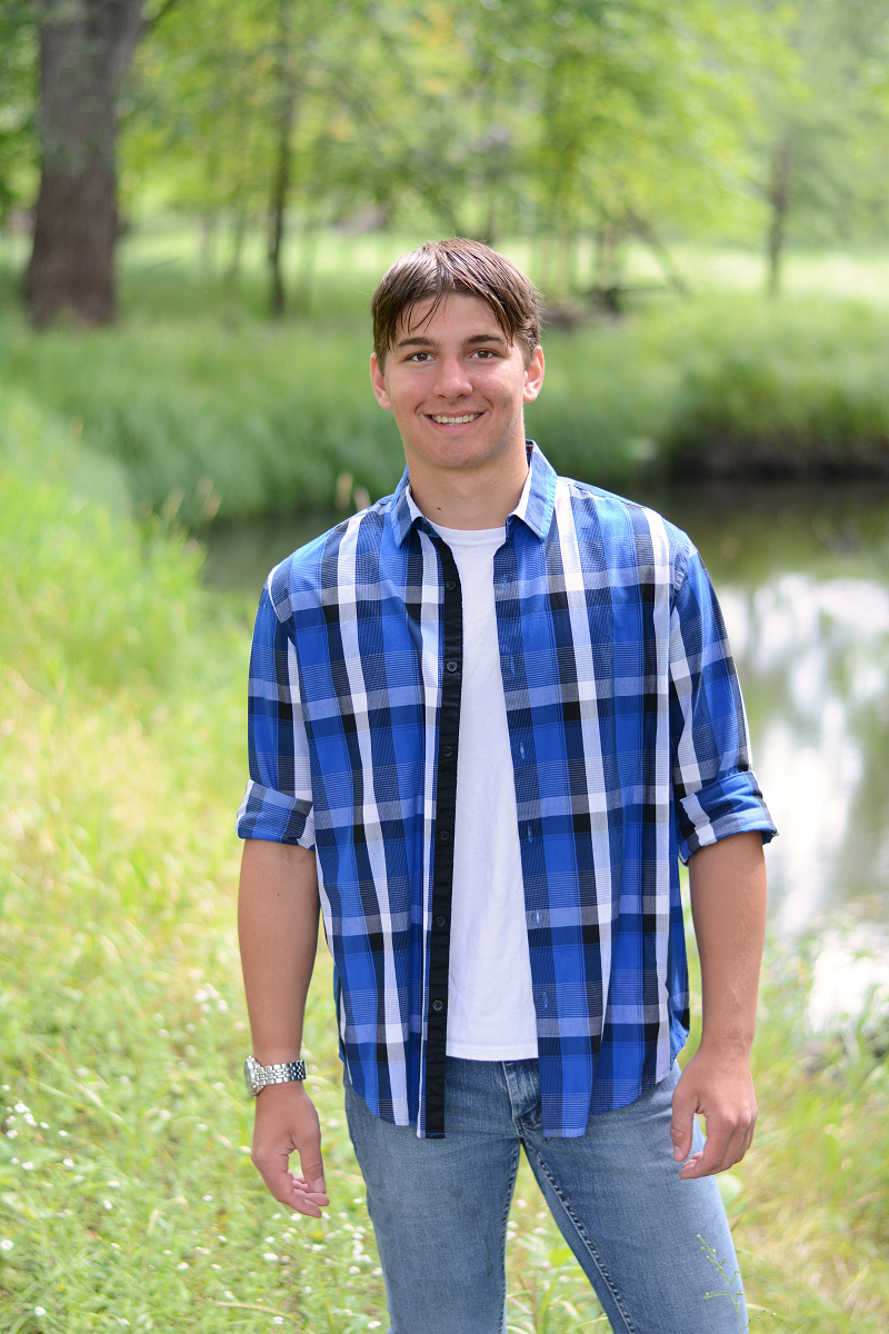 Young man smiling with brown hair wearing a blue flannel shirt with jeans standing in a field of green grass.