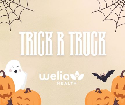 Orange background white text saying "Trick R Truck" with spider webs on upper corners and pumpkins, ghost, and bat on bottom corners. Welia Health white logo centered at bottom.