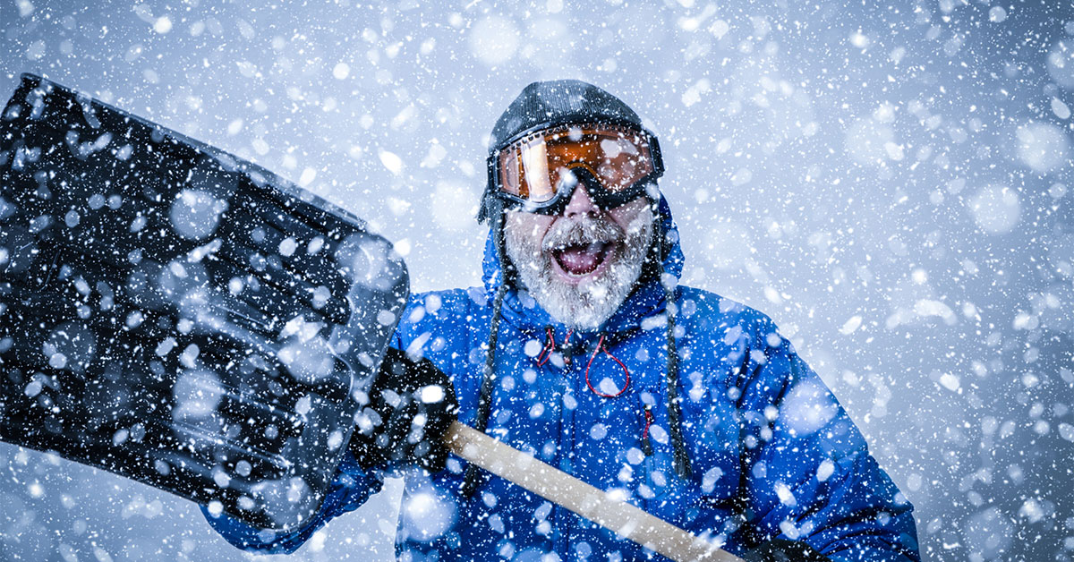 Man bundled in blue parka and snow goggles holds a shovel during heavy snowfall