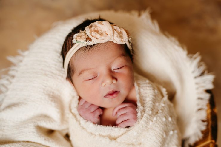 A close up of a newborn baby wrapped in a cream colored blanket sitting upright in a wooden bucket. The baby has dark black hair and is wearing a headband made of peach colored flowers. Her eyes are closed and hands are close to its face.