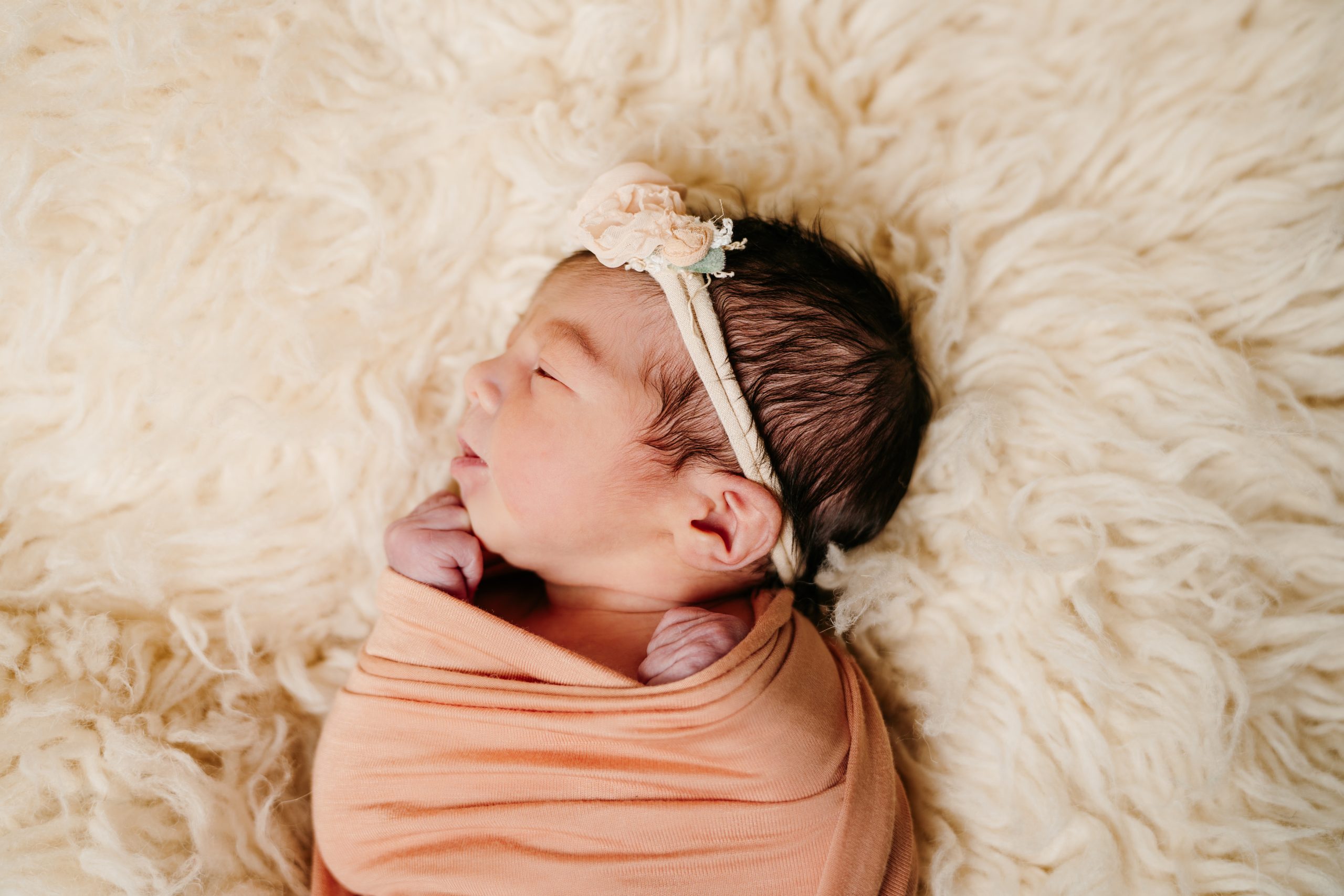 Newborn baby wrapped in a peach colored blanket lying on its side. The baby has dark black hair and is wearing a headband made of peach colored flowers. Her eyes are closed and hands are close to its face.