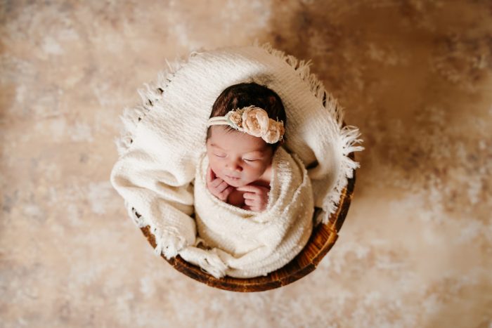 Newborn baby wrapped in a cream colored blanket sitting upright in a wooden bucket. The baby has dark black hair and is wearing a headband made of peach colored flowers. Her eyes are closed and hands are close to its face.
