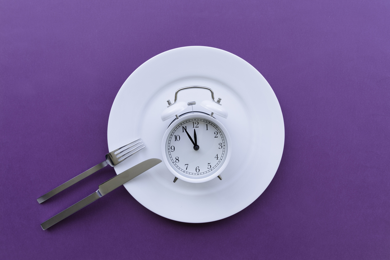 Directly above view of white plate with alarm clock and fork and knife on purple background