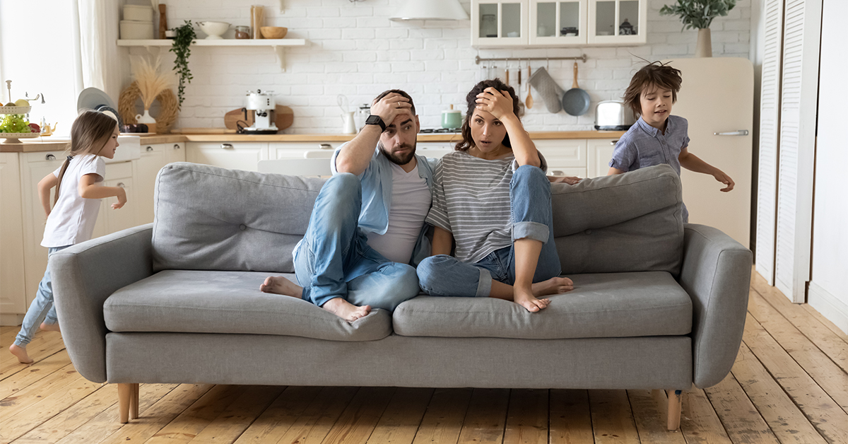 Stressed out parents sitting on a couch as two children run about
