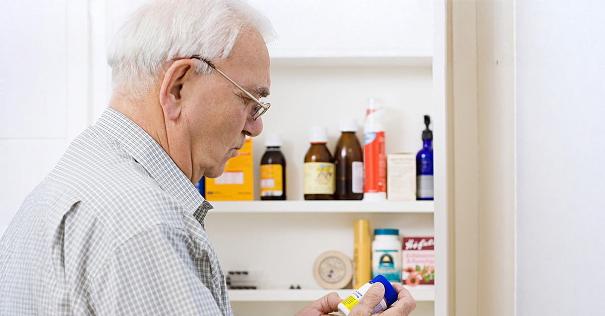 Senior man standing in front of an open medicine cabinet looking at a prescription bottle