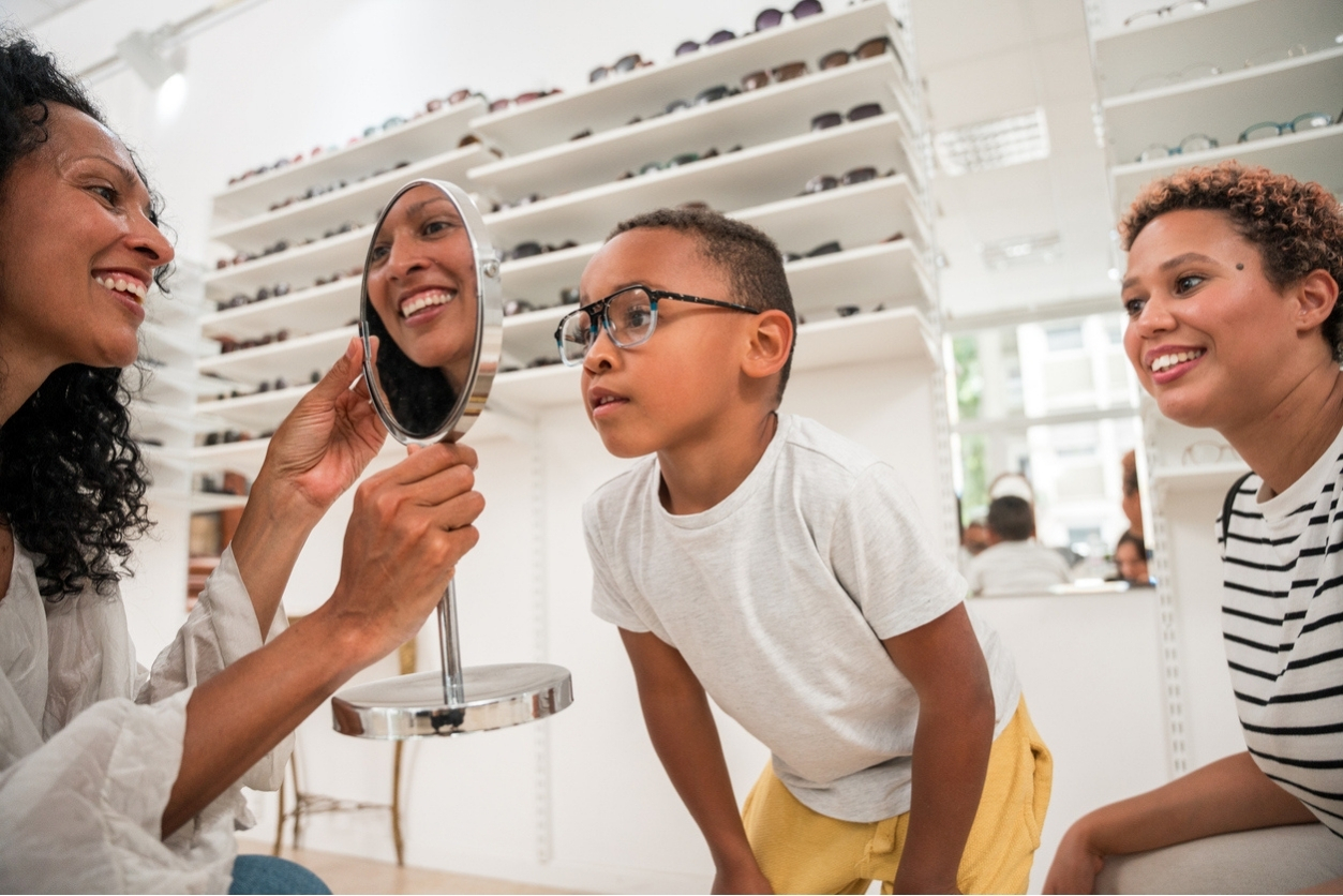 Young boy trying on glasses and looking in the mirror with his mother